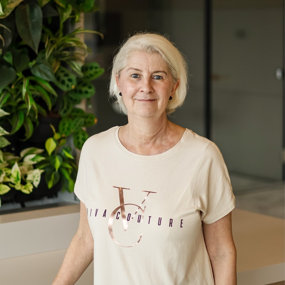 KI-basierte Bildbeschreibung | Eine ältere Frau mit kurzen grauen Haaren steht in einem Innenraum in der Nähe einer vertikalen Pflanzenauslage. Sie trägt ein beigefarbenes Viva Couture-T-Shirt und kleine schwarze Ohrringe und schaut in die Kamera.
