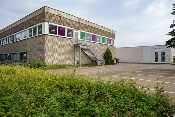 KI-basierte Bildbeschreibung | Ein zweistöckiges, rechteckiges Gebäude mit großen Fenstern und einer Außentreppe neben einem leeren Parkplatz und einigen überwucherten Sträuchern.